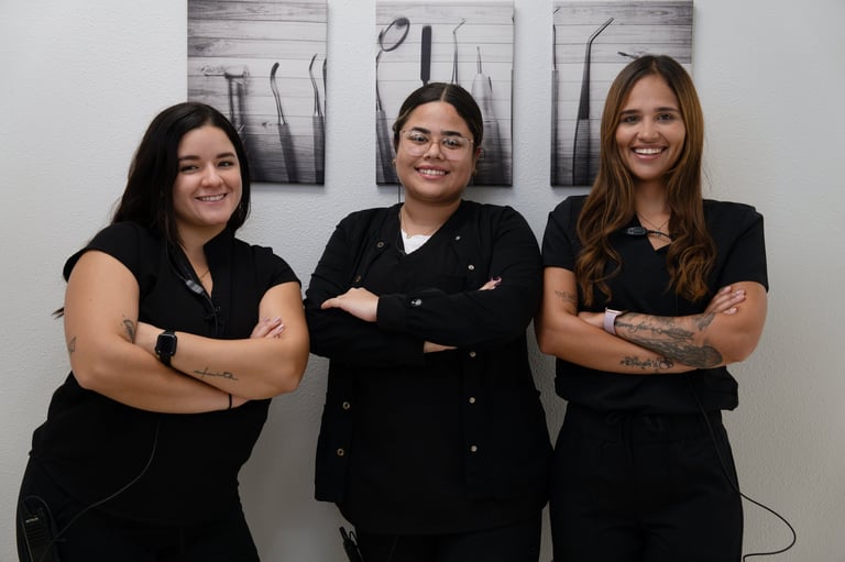 Three women in black outfits stand shoulder-to-shoulder with arms crossed, smiling at the camera in front of a white wall with abstract artwork.